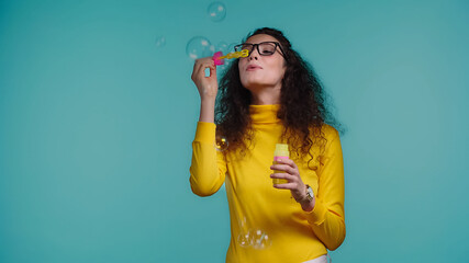 brunette woman in glasses blowing soap bubbles isolated on blue