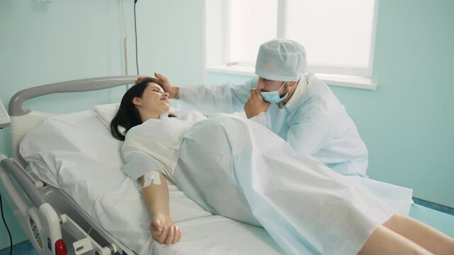 Pregnant woman with painful contractions holding hand of her caring husband that sitting near at hospital ward. Young couple preparing for birth of their child
