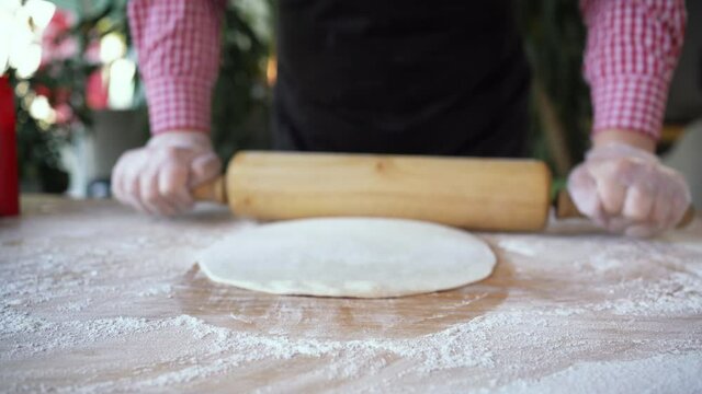 Female chef prepares pizza dough by rolling out with hands and rolling pin on floured table background. Baker with dough in a pizzeria hands close-up