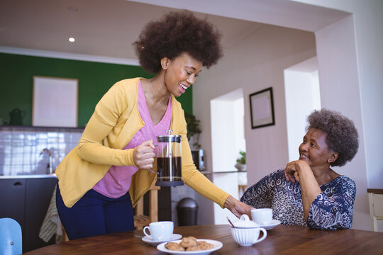 Smiling african american senior woman with adult daughter drinking coffee in kitchen - Powered by Adobe