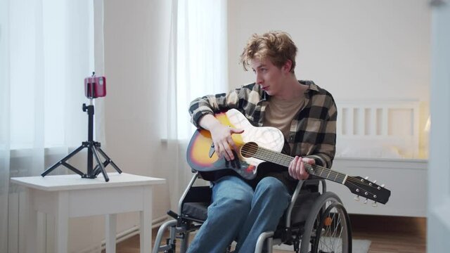 A disabled man is making a video on a phone and playing the guitar