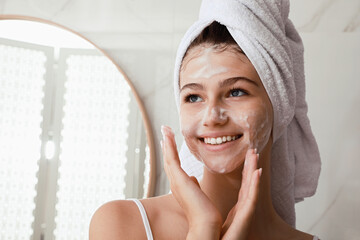 Beautiful teenage girl applying cleansing foam onto face in bathroom, space for text. Skin care cosmetic