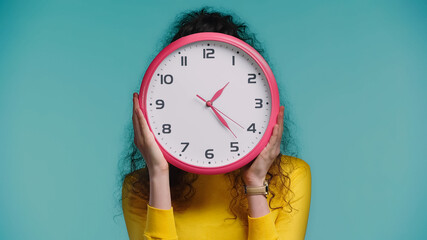 woman obscuring face with wall clock isolated on blue