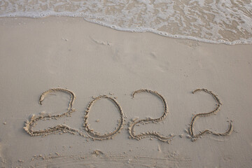Figures 2022 written in white sand of a beach and oncoming foam sea wave. Celebrating the New Year in hot countries. New Year and travel content.