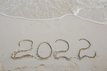 Figures 2022 written in white sand of a beach and oncoming foam sea wave. Celebrating the New Year in hot countries. New Year and travel content.