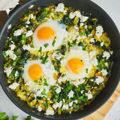 Closeup of classic tasty mediterranean green shakshouka fried with eggs, spinach, celery, parsley, feta cheese and pepper. Served in pan on wooden cutting board.