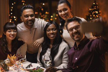 holidays, party and celebration concept - multiethnic group of happy friends having christmas dinner at home and taking selfie with smartphone and waving hands