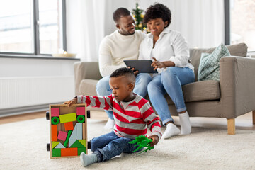 family, winter holidays and people concept - happy african american mother and father using tablet pc computer and baby son playing with toy blocks and dinosaur at home on christmas