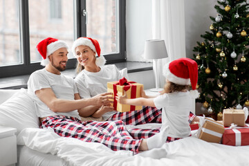 Fototapeta premium family, winter holidays and people concept - happy mother, father and little daughter with christmas gifts in bed at home