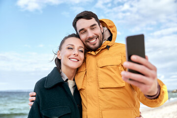 Naklejka premium technology, relationships and people concept - happy couple with smartphone on autumn beach