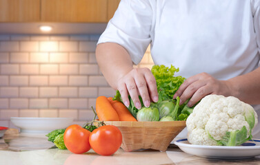 Close up of overweight woman hand preparing various organic vegetables into bamboo basket for cooking on kitchen table, healthy lifestyle concept
