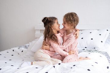 Obraz na płótnie Canvas two beautiful diverse kids girls in pajamas hugging on bed in modern bright apartment