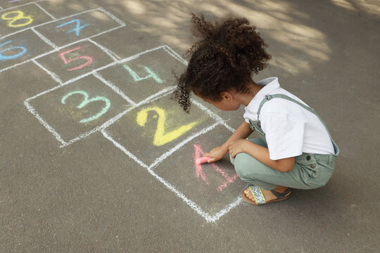 Little African American girl drawing hopscotch with chalk on asphalt outdoors. Happy childhood