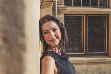 Dark-haired woman posing in a black dress by the wall with window. The woman is wearing black sunglasses. It's a beautiful sunny day.