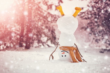 Cheerful snowman with orange scarf and in yellow boots stands upside down in winter forest