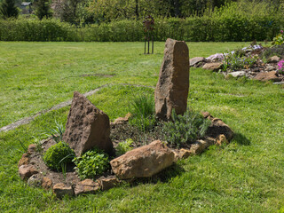 View on spring garden with oval flower bed with green herbs thyme, lavender, mint, chive and sweet balm with big sandstone rock and stones on background of lush green grass