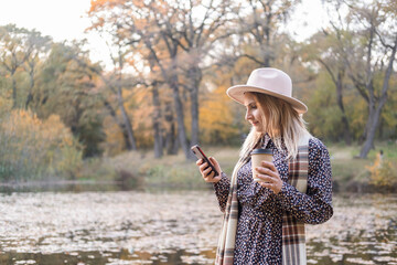 beautiful young woman drinking coffee, using phone in nature in autumn park in fall