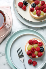 Delicious tartlets with berries and tea on white marble table, flat lay