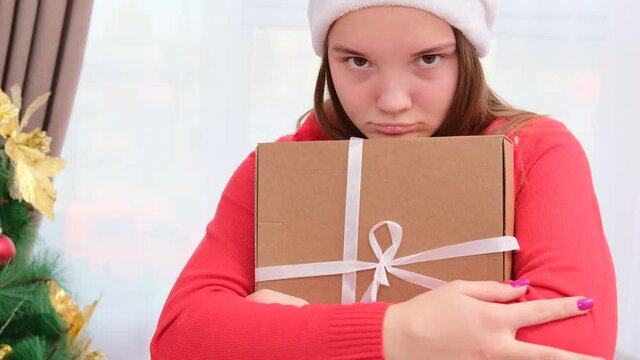 Close up video of a girl in a Santa hat with sad facial expression holding the gift and does not want to give yo anyone.