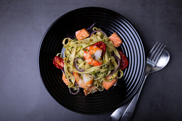 Noodles with seafood, sun-dried tomatoes, capers and red onions. Homemade pasta with shrimp, salmon (trout) and pesto sauce. Black background, black plate. Close-up.
