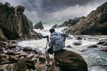 Photographer man holding with camera, tripod and facing the rainstorm on seashore