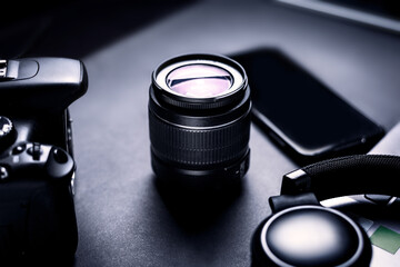 Designer's workspace. Camera lens on a dark table with camera body, headset, laptop and smartphone.
