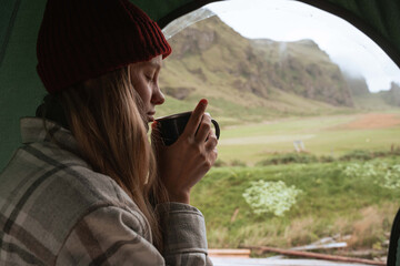 Woman sitting inside the tent at the camping and drinking hot coffee while started her day
