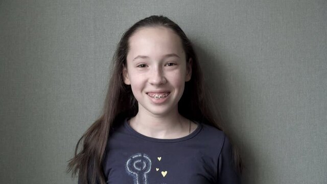 A lovely young teenage girl with braces on her teeth looks into the camera and smiles broadly, standing at home by the window against a green wall. A girl with long hair in casual clothes. Close-up.