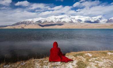 A woman in red cloak sits by the lake and looks at the Muztagh Ata peak.