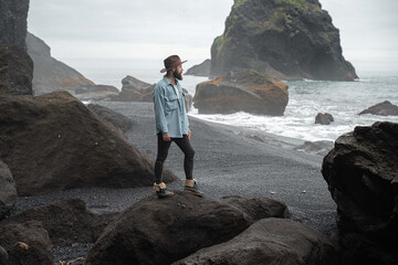 Bearded man looking at the wavy ocean while spending time at the location with basalt sand