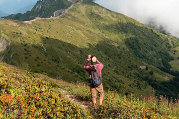 Naklejka premium Young man with backpack hiking in Caucasus green mountains among bushes of rhododendron and taking photo on smartphone, using modern technology active and healthy lifestyle, landscape
