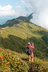 Young man with backpack hiking in Caucasus green mountains among bushes of rhododendron and taking photo on smartphone, using modern technology active and healthy lifestyle, landscape