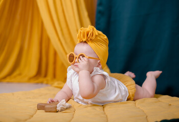 A fashionable baby girl is resting in yellow glasses. Cute girl posing in the studio in a yellow beanie cap