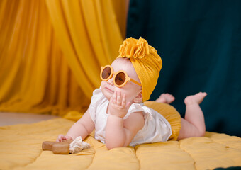 A fashionable baby girl is resting in yellow glasses. Cute girl posing in the studio in a yellow beanie cap