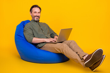 Portrait of attractive cheery man sitting in bag chair using laptop typing project isolated over bright yellow color background