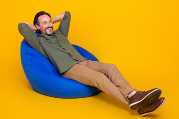 Portrait of attractive cheerful dreamy man resting in bag chair free time isolated over bright yellow color background