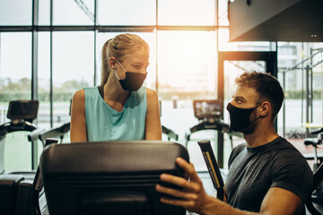 Young woman at body workout in modern gym together with her personal fitness instructor or coach. They keeping distance and wearing protective face masks. Coronavirus world pandemic and sport theme.
