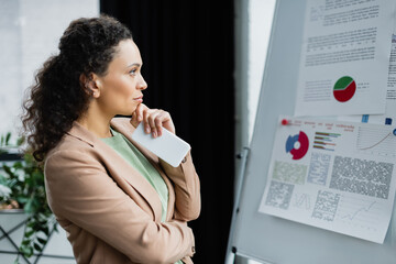 Fototapeta na wymiar thoughtful african american businesswoman with mobile phone looking at flip chart with analytics in office