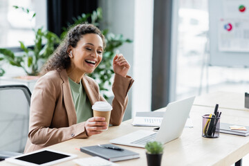 african american businesswoman with coffee to go smiling during video call on laptop in office