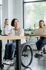 thoughtful businesswoman in wheelchair holding takeaway drink near blurred african american colleague in office