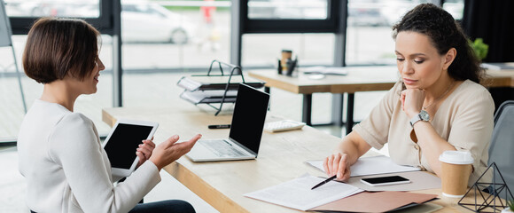 positive businesswoman pointing with hand near thoughtful african american colleague working with documents, banner