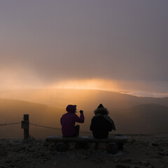 two girls are sitting on a bench on the top of the mountain, meeting the dawn in autumn and...