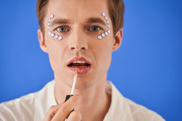 Man with rhinestones under eyes applying lip gloss and looking attentively at the camera