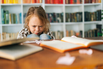 Front view of tearing upset primary child school girl sitting alone hugging knees in front of desk with difficult homework. Sad schoolgirl put head on knee being stressed with exam, selective focus