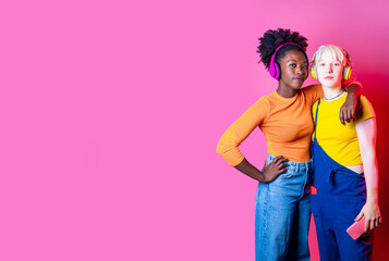 Two young diverse multicultural women best friends posing isolated background having fun laughing holding smartphone wearing wireless headphones