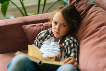 High-angle view of sad adorable child kid girl reading paper book lying on soft couch at home looking at camera. Top view of little cute upset girl lying on sofa alone in cozy living room.
