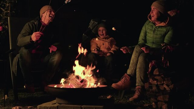 Happy family of mom dad and their son roast marshmallows on a fire pit their backyard garden