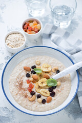 Plate of oatmeal porridge with dried banana, candied fruits and nuts, vertical shot on a white concrete background