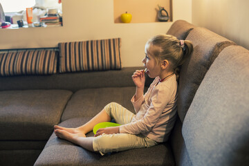 Obraz premium Beautiful little girl eating unhealthy food while watching TV at the sofa