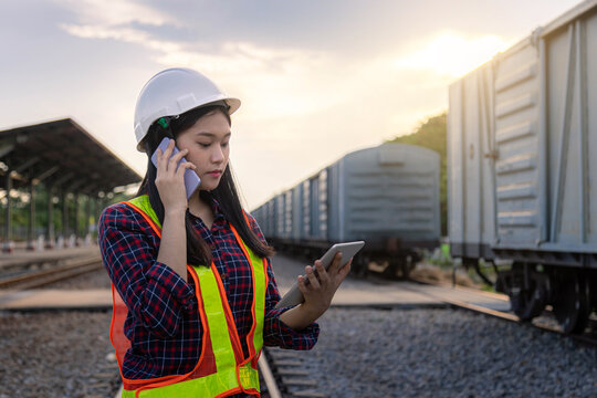 Women engineer railway wearing safety uniform holding tablet and talking telephone on hand for inspection and check container truck railway distribution.